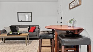 Una sala de estar moderna con un sofá gris, una mesa de centro de madera y una mesa de comedor redonda con dos taburetes de cuero negro.