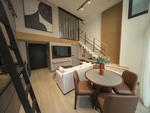 Ein modernes Wohnzimmer mit einer Sofagarnitur, einem Esstisch mit Stühlen, einem Fernseher und einer Treppe.