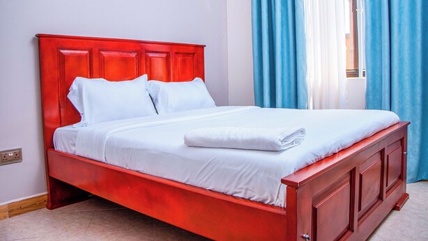 A red bed with white linens, a wooden headboard, and a window with blue curtains. - Gugga Suites Mbale (Mbale)