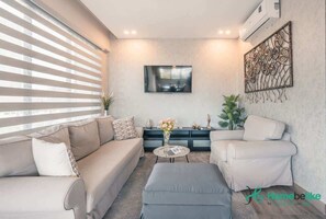 A modern living room with a beige sofa, a grey ottoman, and a white armchair. There is a flat-screen TV mounted on the wall, a decorative metal piece, and a small table with a vase of flowers. - Regatta Living Ii with Breakfast Included! - 502 (Santo Domingo)