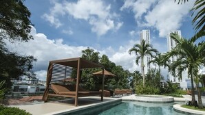 Una piscina con una pérgola y sillas de descanso, rodeada de palmeras y edificios modernos.