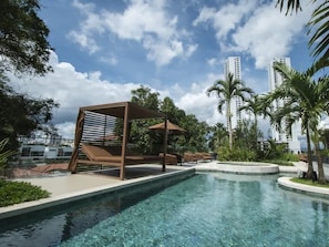 Ein Swimmingpool mit einer Pergola und Liegestühlen, umgeben von Palmen und modernen Gebäuden.