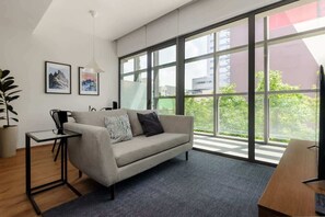 A modern living room with a sofa, a small table, and a large window offering a view of greenery and buildings. - Cozy Furnished Studio in Mexico City U203 (Ciudad de México)