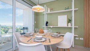 A dining area with a round table set for four, white chairs, a hanging light fixture, and a view of greenery outside. - Stunning 1BR Apt with amazing City Views (Asunción)