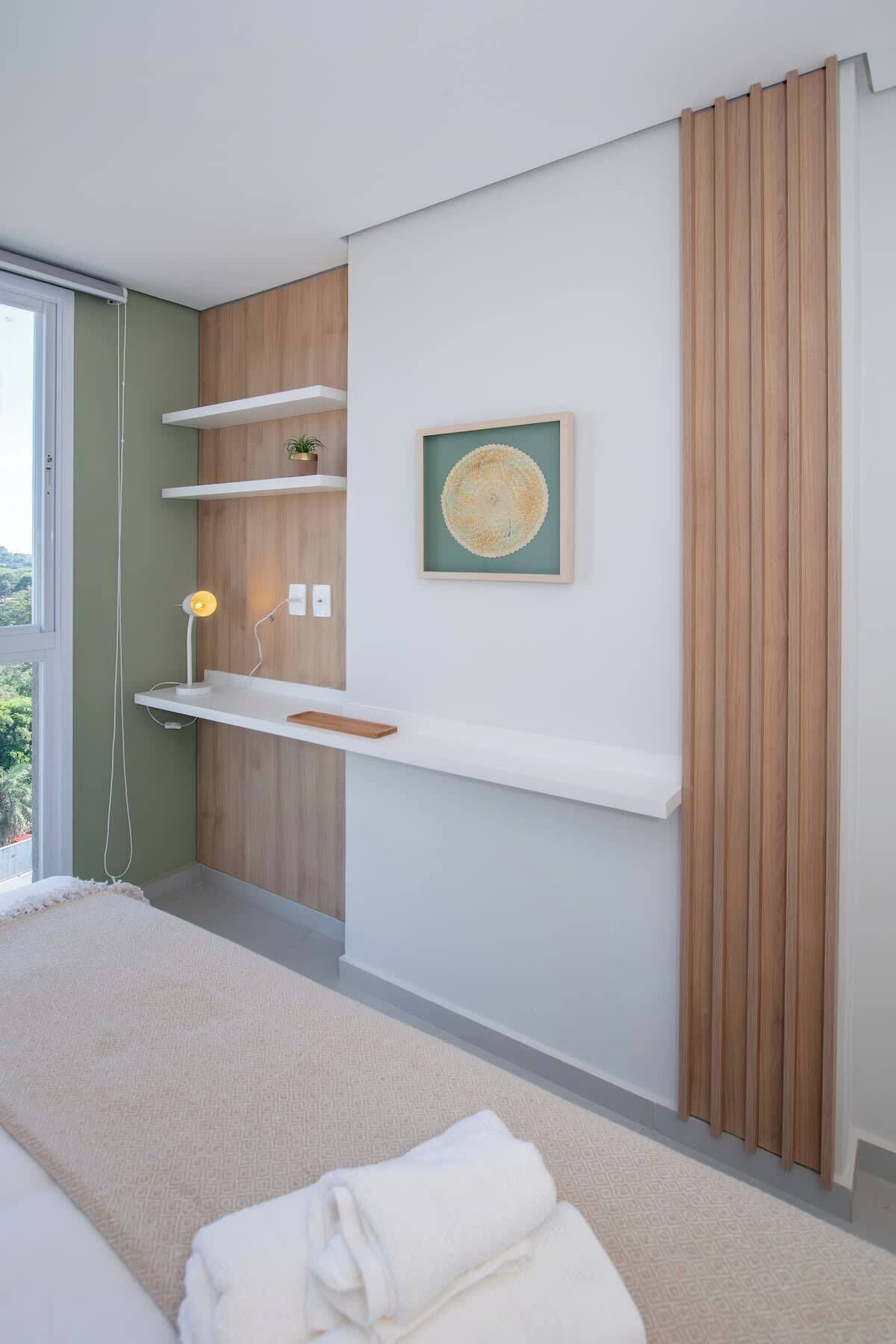 A modern bedroom with a bed, a bedside table, a wall-mounted shelf, a framed artwork, and a window with a view.