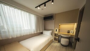 Een compacte hotelkamer met een enkel bed, een klein nachtkastje, een inbouwbankje, een raam met dunne gordijnen, een wandgemonteerde airconditioning, een bedlampje en een klein wastafeltje met spiegel.
