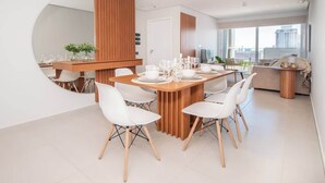 Un comedor moderno con una mesa de madera y sillas blancas, una zona de cocina con mostrador de madera, y un amplio ventanal con vistas a la ciudad.