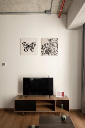A living room with a flat-screen TV on a wooden stand, two wall-mounted artworks, a small table with a vase, and a window with curtains. - Furnished Studio in Mexico City (Ciudad de México)