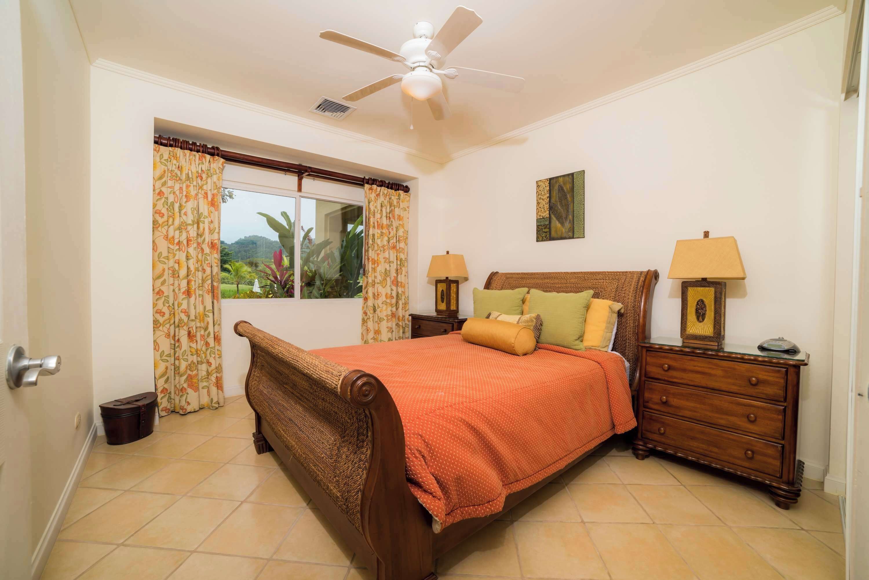 A bedroom with a bed, two bedside tables with lamps, a ceiling fan, and a window with curtains.