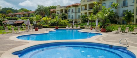 Une grande piscine extérieure à plusieurs niveaux, entourée de bâtiments et bordée d’une végétation luxuriante et de palmiers.