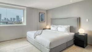 A bedroom with a large bed, a nightstand with a lamp, a framed picture on the wall, and a view of the city through a large window. - Modern King Studio in Central Roma Norte (Ciudad de México)