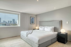 A bedroom with a large bed, a nightstand with a lamp, a framed picture on the wall, and a view of the city through a large window. - Modern King Studio in Central Roma Norte (Ciudad de México)