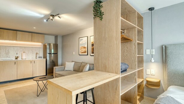A modern kitchen with wooden cabinets, a dining area with a bar stool, a sofa, and a built-in wooden shelving unit with a hanging plant. - Modern King Studio in Central Roma Norte (Ciudad de México)