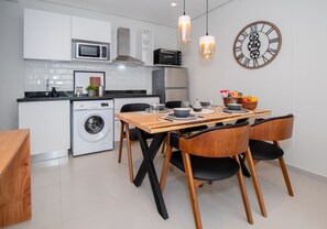 A compact kitchen with a microwave, toaster, and washing machine, a wooden dining table with chairs, and a wall clock. - Sleek 1BR Apt, City Views & Pool (Asunción)