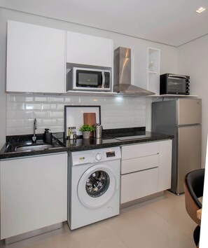 Eine kompakte Küche mit weißen Schränken, einer eingebauten Mikrowelle, einem Toaster, einem Edelstahlkühlschrank und einer Frontlader-Waschmaschine.