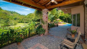 Un patio cubierto con ventilador de techo, piso de piedra, plantas en macetas, piscina y vistas a un paisaje exuberante.
