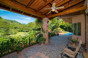 A covered patio with a ceiling fan, stone flooring, potted plants, a pool, and a view of a lush landscape. - Casa Oasis 5 bdr private pool (Playa Herradura)