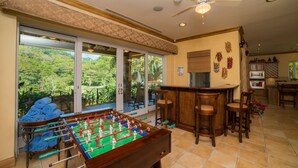 Una habitación con mesa de fútbolín, zona de bar y amplios ventanales con vistas a un paisaje exuberante.