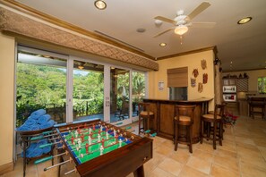 A room with a foosball table, bar area, and large windows overlooking a lush landscape. - Casa Oasis 5 bdr private pool (Playa Herradura)
