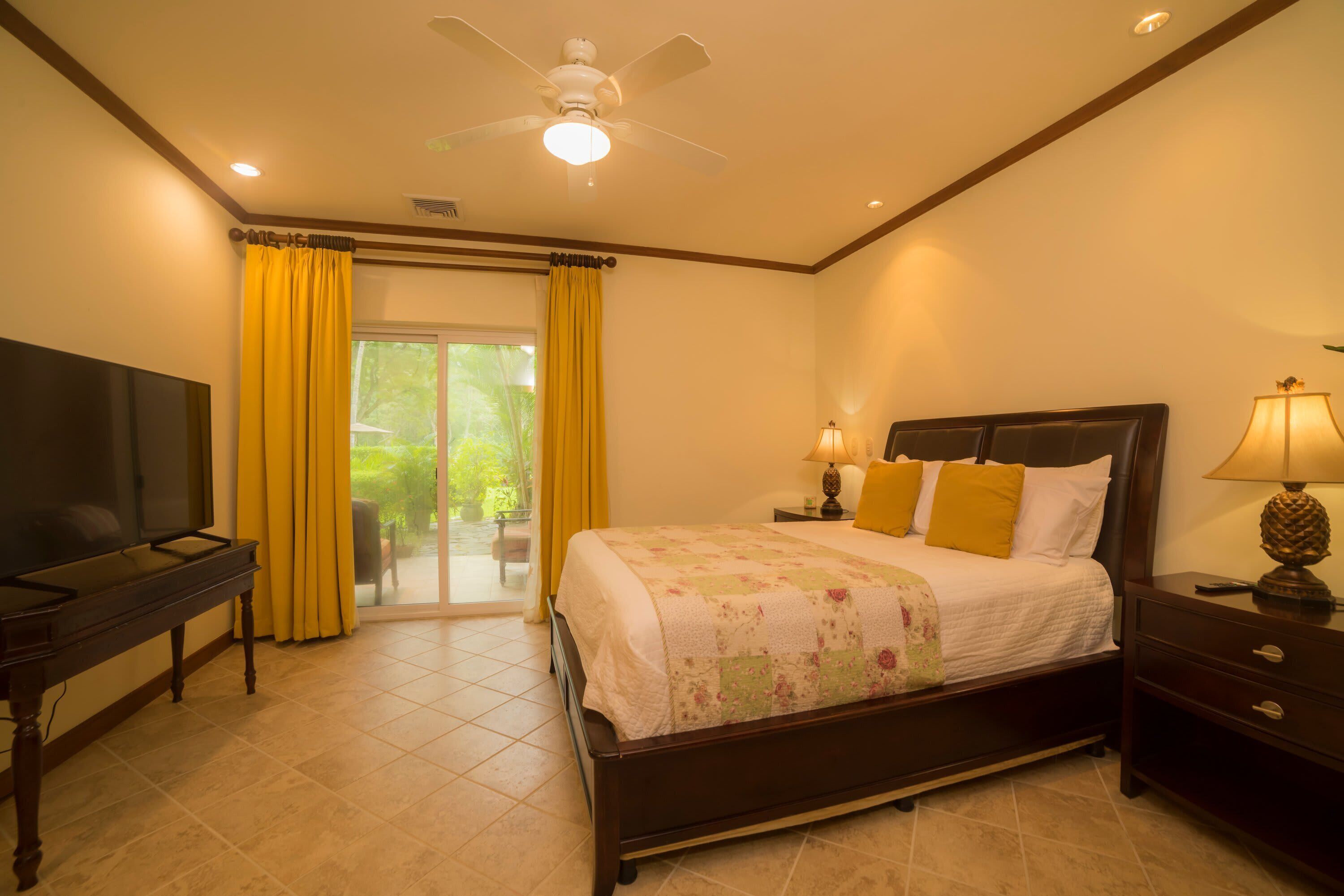 A hotel room with a bed, a television, a ceiling fan, and a view of the outdoors.