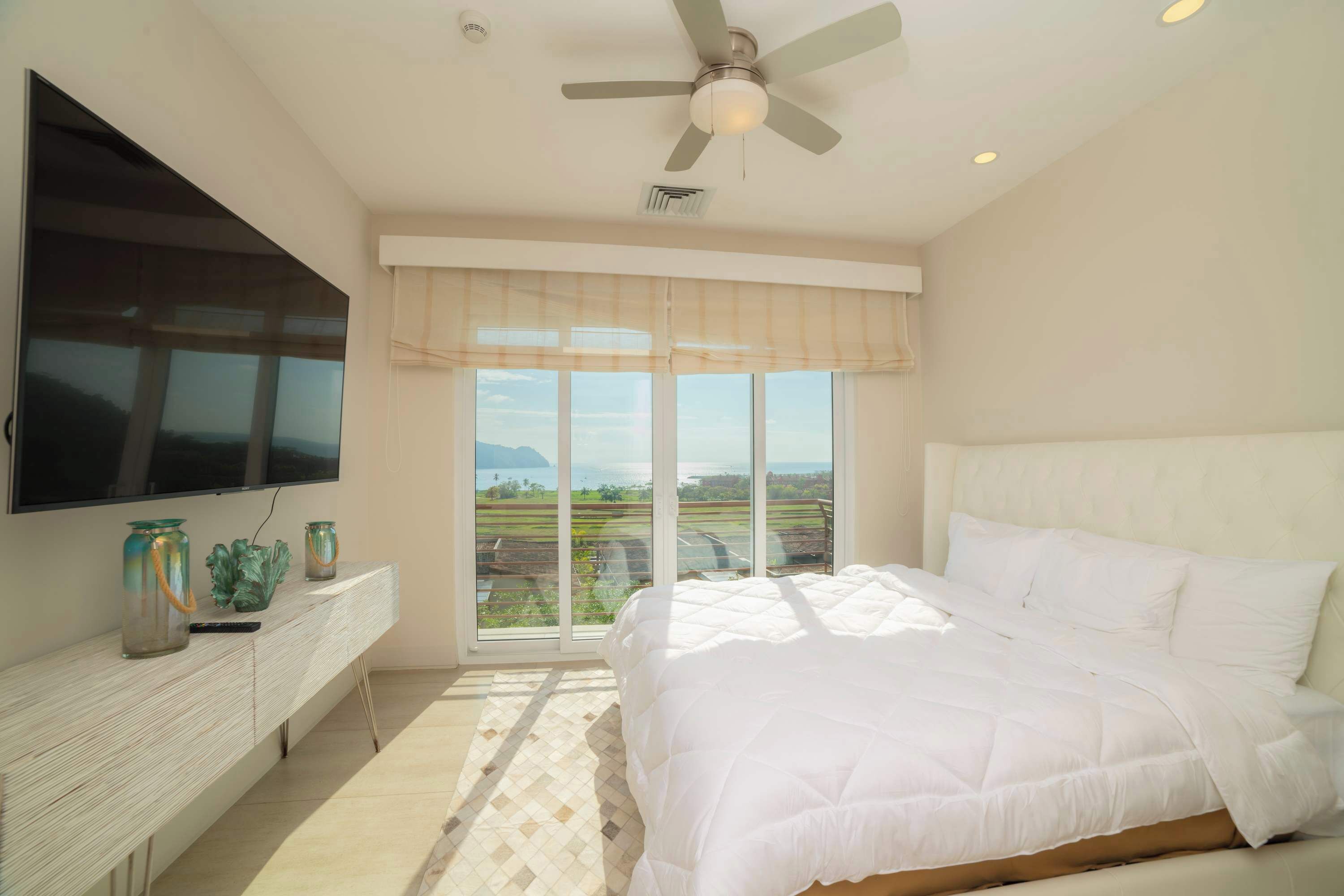 A bedroom with a bed, a TV mounted on the wall, a ceiling fan, and a large window with a view.