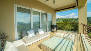 Un balcone con divano, tavolino in vetro e vista su vegetazione e colline.
