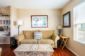 A living room with a beige sofa, a coffee table, a bookshelf, a framed picture, and a window with blinds. - Gulf Views, Pet Friendly, Deck (Bolivar Peninsula)