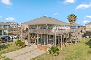 A two-story wooden house with a balcony and a deck, surrounded by grass and a palm tree. - Gulf Views, Pet Friendly, Deck (Bolivar Peninsula)