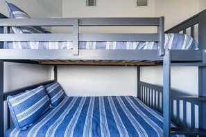 A bunk bed with a striped bedspread and pillows, a window with blinds, and a ventilation grille on the wall.