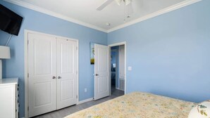 Un dormitorio con una cama, dos puertas blancas, un ventilador de techo y un televisor montado en la pared.