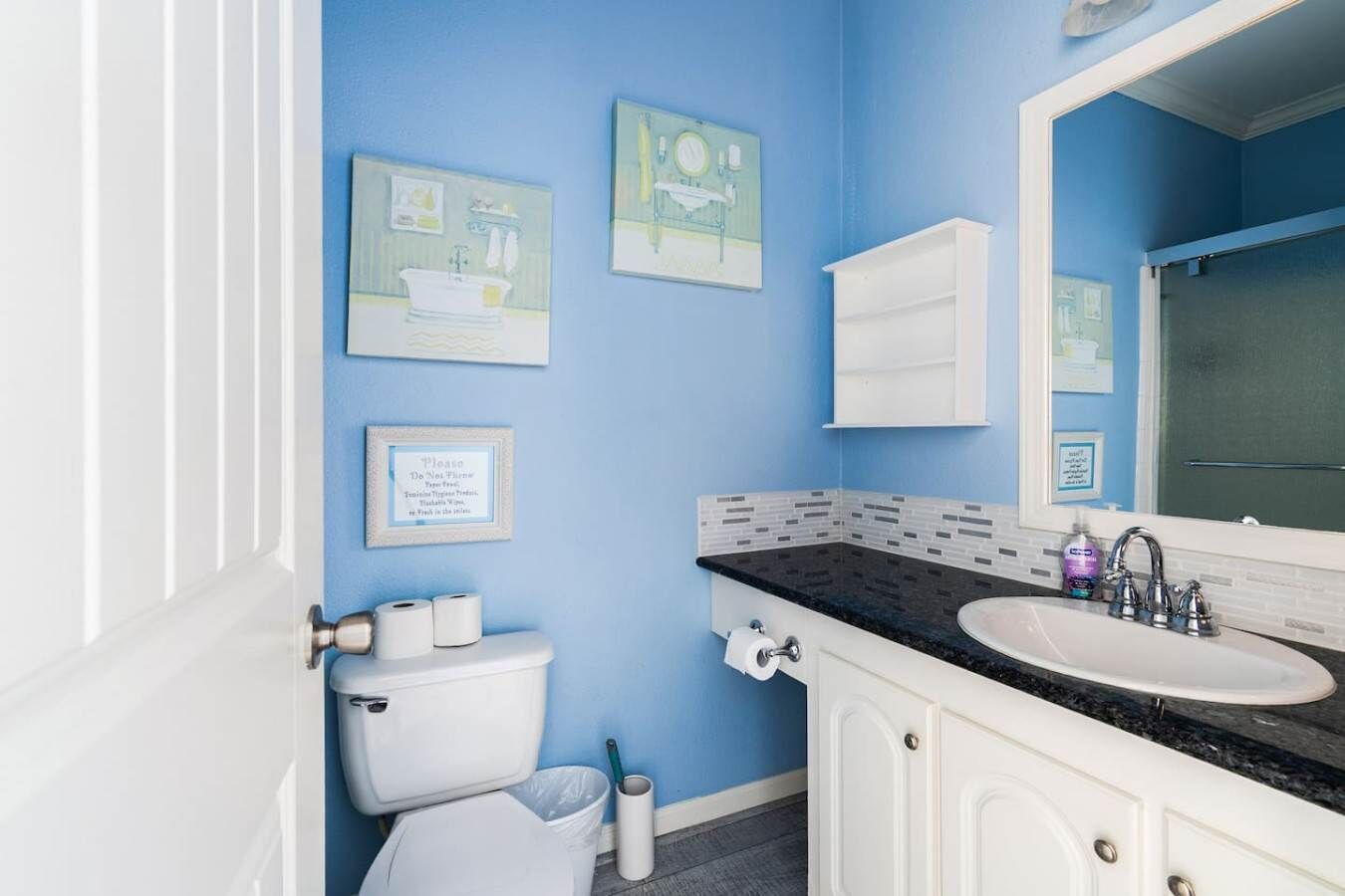 Ein Badezimmer mit Toilette, Waschbecken und Spiegel. An den Wänden hängen Bilder in Rahmen und ein Mülleimer steht bereit.