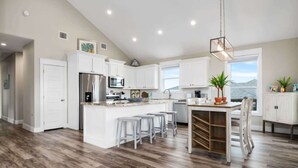 A modern kitchen with a central island, white cabinetry, and a dining area with bar stools. - Two Adjacent Cabins, Sleeps 18 (Bolivar Peninsula)