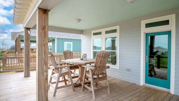 A covered outdoor deck with wooden chairs and a table, a sliding glass door, and a view of a neighboring house. - Two Adjacent Cabins, Sleeps 18 (Bolivar Peninsula)
