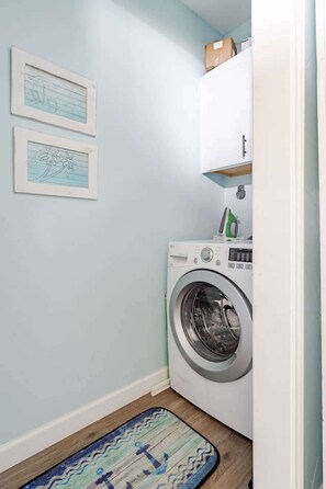 A laundry room with a washing machine, a shelf with items, and two framed pictures on the wall. - Fire Pit, Playscape, The Red Mermaid Retreat (Bolivar Peninsula)