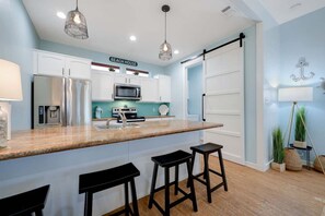 A modern kitchen with white cabinets, a stainless steel refrigerator, a microwave, and a coffee machine. There are two black bar stools and a large island with a sink. - Fire Pit, Playscape, The Red Mermaid Retreat (Bolivar Peninsula)