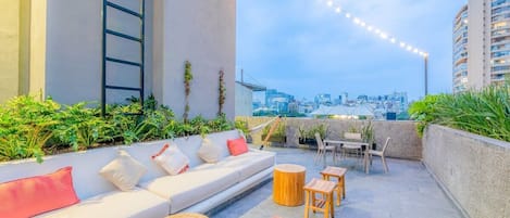 Eine Dachterrasse mit einer Couch, Holztischen und Stühlen mit Blick auf eine Stadtlandschaft.
