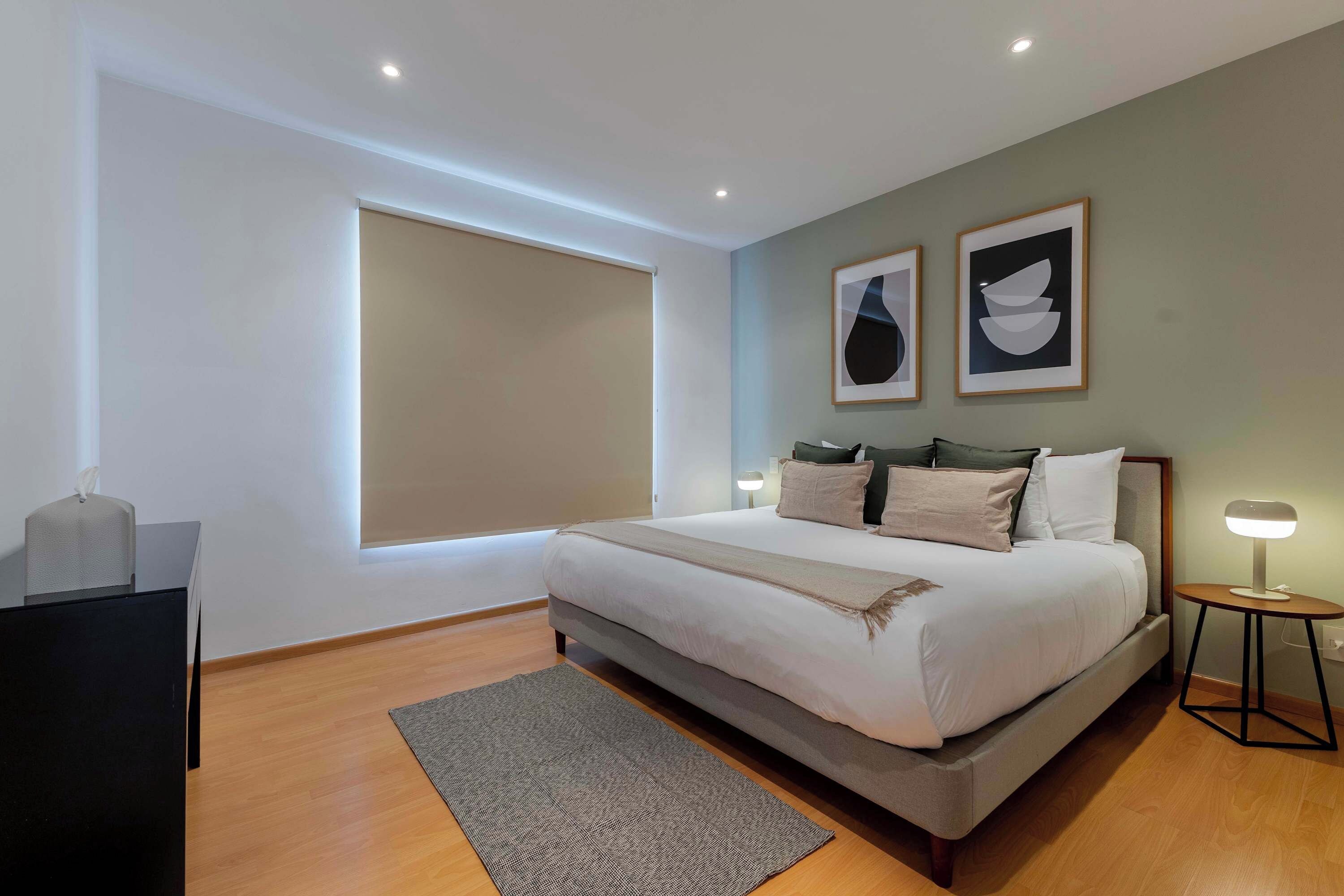 A modern bedroom with a large bed, a bedside table, a lamp, and two framed artworks on the wall.