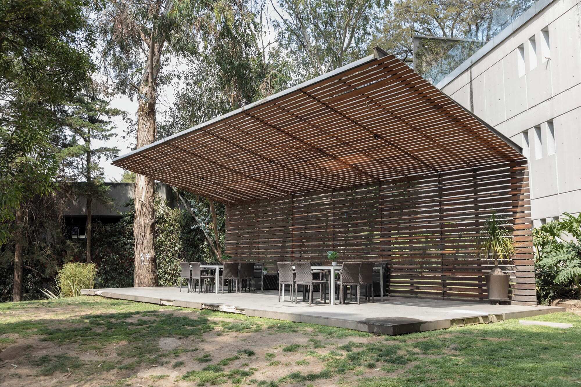 An outdoor seating area with a slatted roof structure, surrounded by greenery and a building in the background.