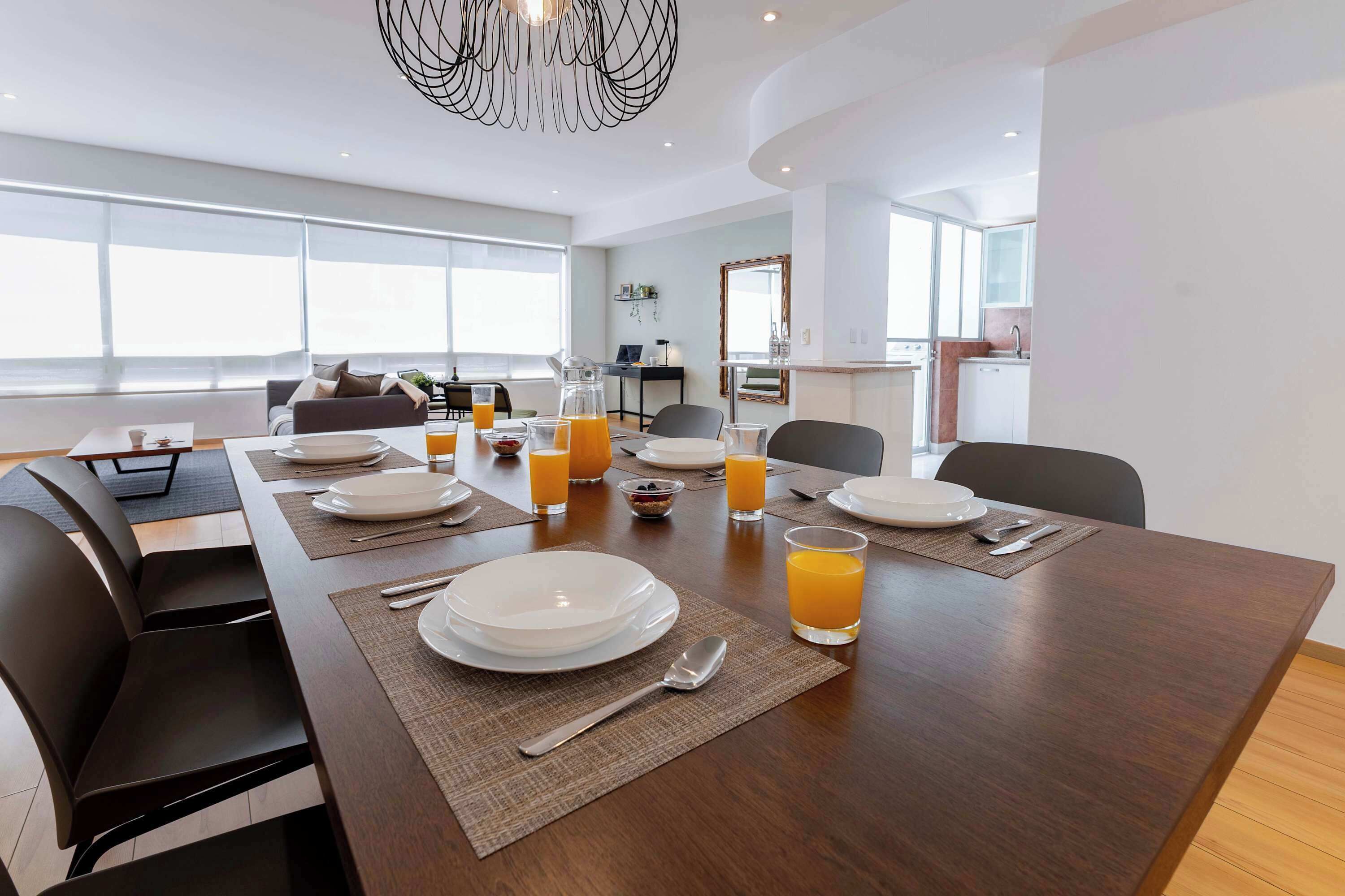 A modern dining area with a long table set for breakfast, including plates, glasses of orange juice, and cutlery.