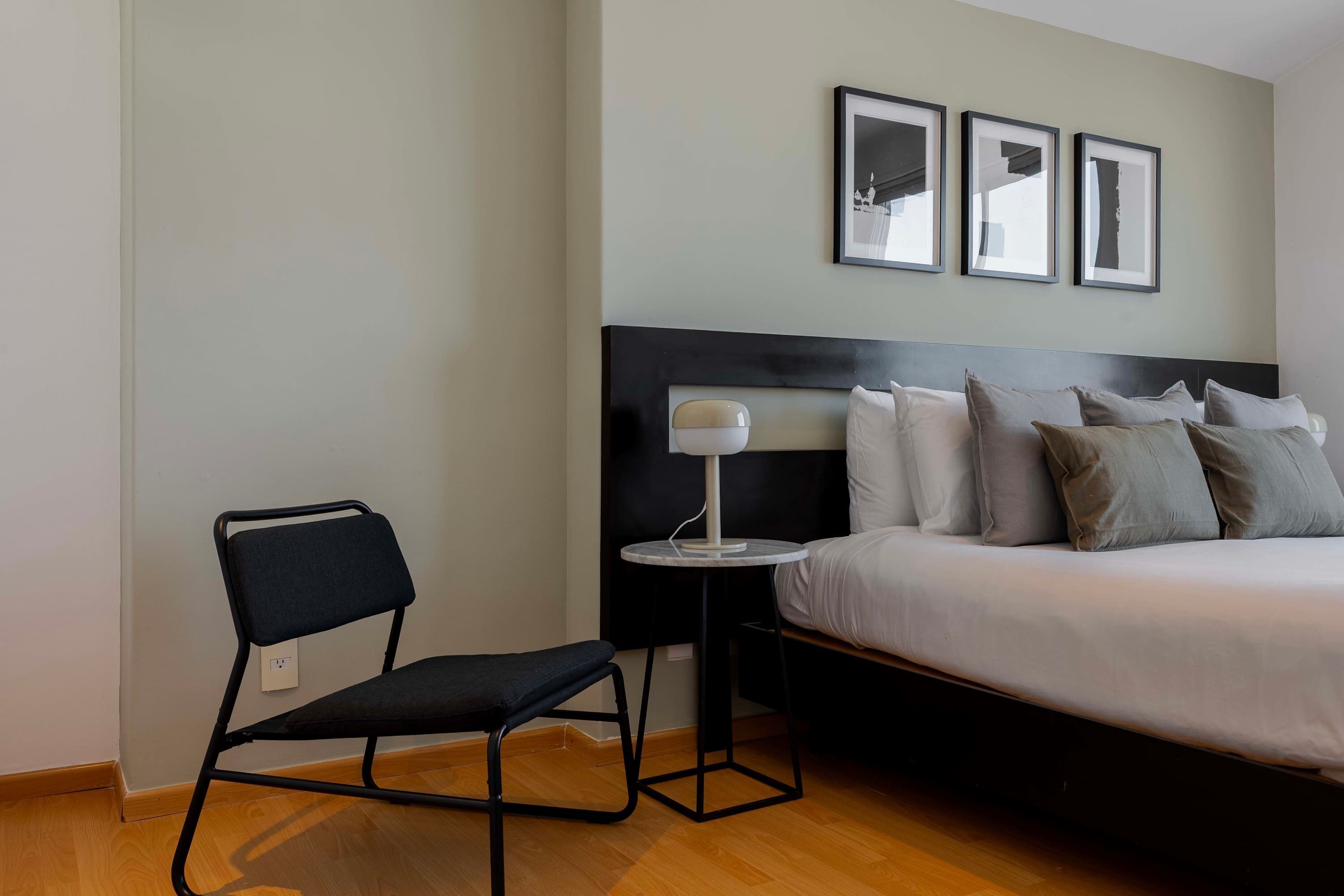A hotel room with a bed, a chair, a small table with a lamp, and three framed pictures on the wall.