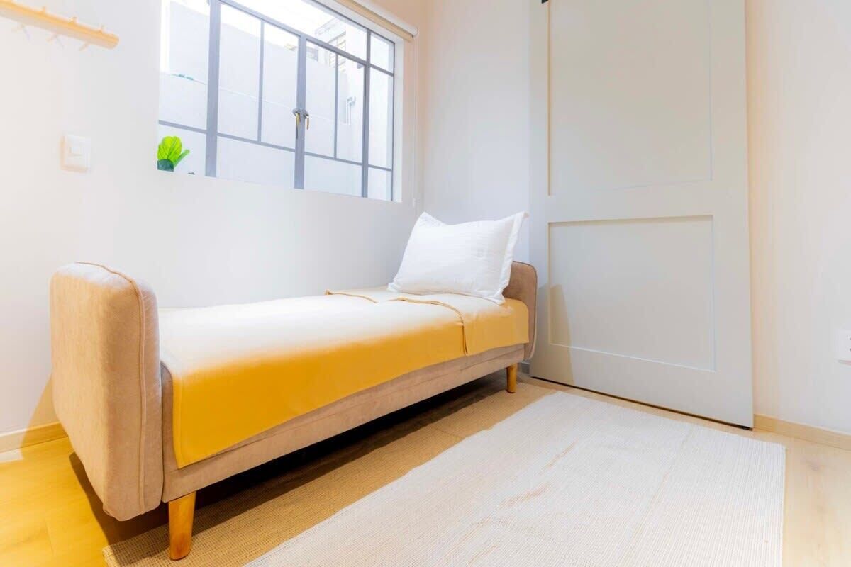 Ein Zimmer mit einem Einzelbett, einem Fenster, einer Tür und einer kleinen Pflanze an der Wand.