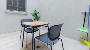 En liten sittplats utomhus med ett runt bord och två stolar, ett fönster, en krukväxt och städredskap.