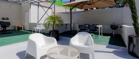 Eine Außenpatio-Terrasse mit weißen Plastikstühlen, einem runden Tisch, einem Grill und einem großen Sonnenschirm.