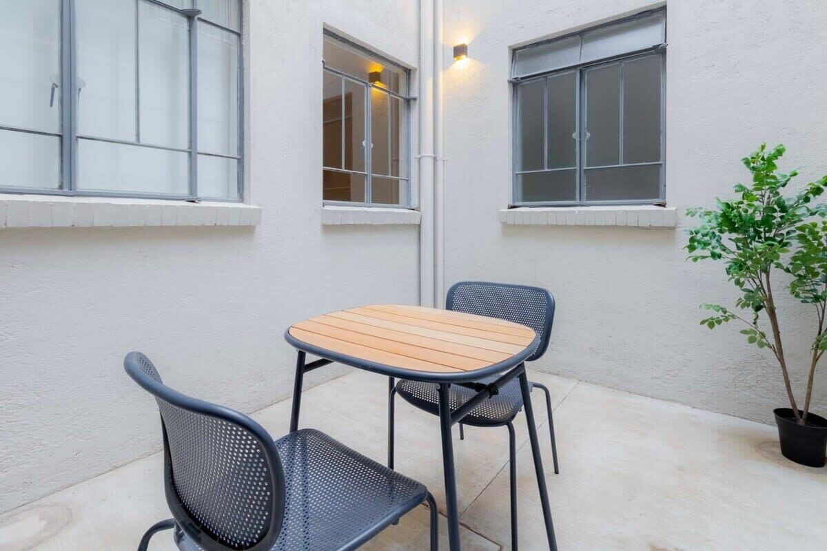 A small patio with a wooden table and two chairs, a potted plant, and a white wall with windows.