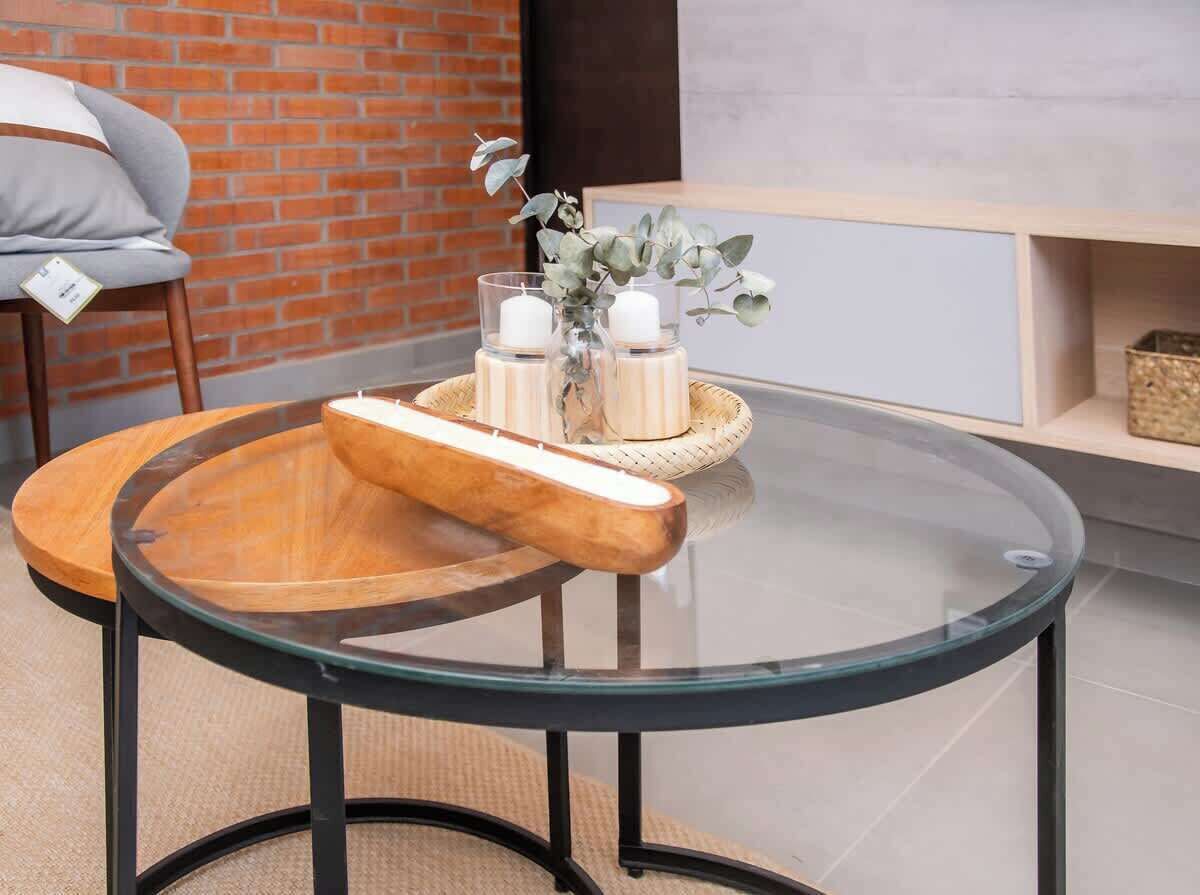 Una mesa redonda de cristal con una bandeja de madera y velas, frente a una pared de ladrillo y un mueble blanco.