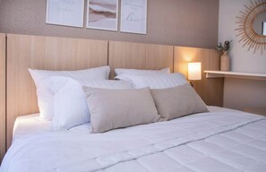 Ein ordentlich bezogenes Bett mit weißer Bettwäsche und beigen Kissen, ein an der Wand befestigter Spiegel und gerahmte Kunstwerke darüber.