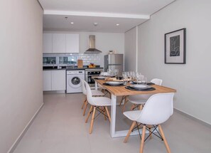 Una cocina moderna con armarios blancos, una estufa de acero inoxidable y un comedor con mesa de madera y sillas blancas.
