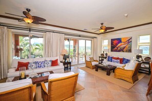 Amplia sala de estar con mobiliario de mimbre, ventilador de techo y grandes ventanales con vistas al exterior.