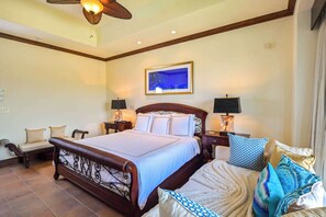 Habitación de hotel con una cama grande, dos mesitas de noche con lámparas, un ventilador de techo, un cuadro enmarcado en la pared y un área de descanso con sofá y cojines.