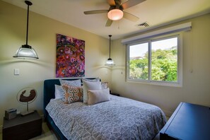 Ein Schlafzimmer mit einem Bett, zwei Pendelleuchten, einem Deckenventilator, einem Fenster mit Ausblick und einem Bild an der Wand.